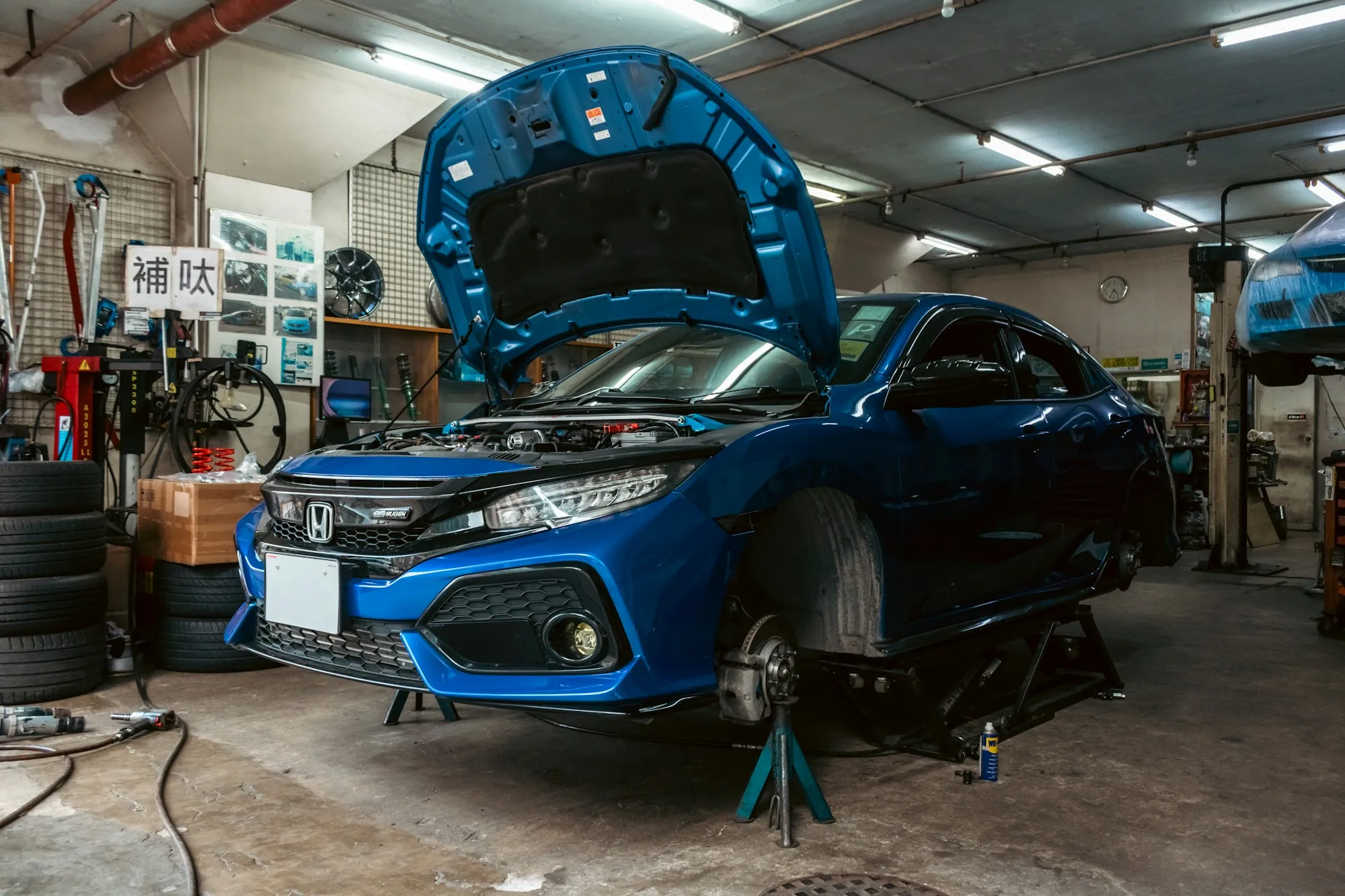 Blue hatchback raised on a lift inside a busy repair shop